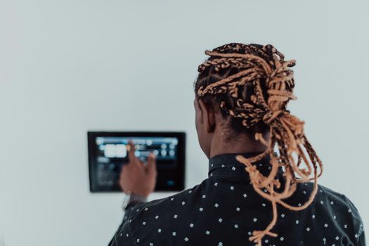 Smiling African American man using modern smart home system, controller on wall, positive young man switching temperature on thermostat or activating security alarm in apartment. High quality photo