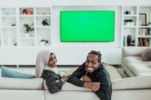 African Couple Sitting On Sofa Watching TV Together Chroma Green Screen Woman Wearing Islamic Hijab Clothes. High quality photo