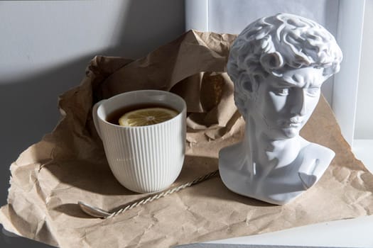 A white corrugated cup with tea and lemon, with a plaster head sculpture of Apollo and a cupronickel teaspoon on a long handle is on brown craft paper on a wooden table.