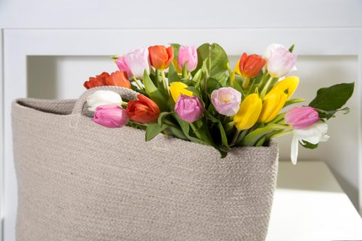 Bouquet of multicolored tulips in a white rattan wicker bag standing on the table in interior.