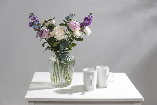 Bouquet of hackelia velutina, purple and white roses, small tea roses, matthiola incana and blue iris in glass vase is on the white coffee table with two tall cups for tea. Grey wall