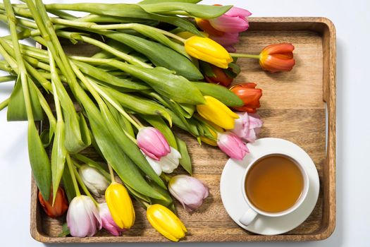 A bouquet of multicolored tulips and a cup of tea are on a wooden tray on the table. Copy space