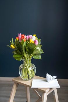 A bouquet of multi-colored tulips in a transparent vase on the tall chair with step against a dark blue background. White notebook.
