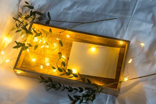 A eucalyptus branch lies on a white sheet beside a tray with a letter in an envelope. The garland is glowing. Copy space. Place for text.