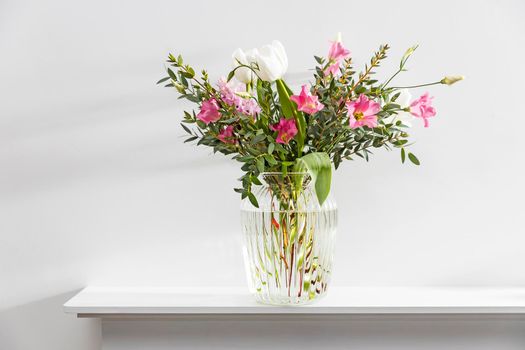 A minimalistic bouquet of white tulips, pink eustoma, hyacinth, eucalyptus in a fluted glass vase on a white panel of an artificial fireplace. Empty space.