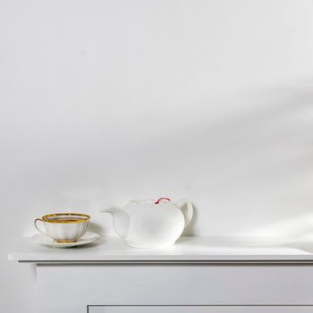 White on white. A cup of tea, a kettle, and a notebook stand on a white panel of an artificial fireplace. Empty space.