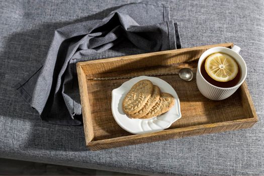 A white cup of tea with lemon, a long cupronickel spoon with a twisted handle and a saucer with three oatmeal cookies for breakfast on a wooden tray, a rag napkin on a gray sofa. Serving