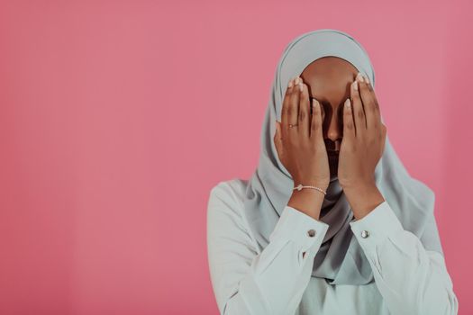 Modern African Muslim woman makes traditional prayer to God, keeps hands in praying gesture, wears traditional white clothes, has serious facial expression, isolated over plastic pink background. High-quality photo