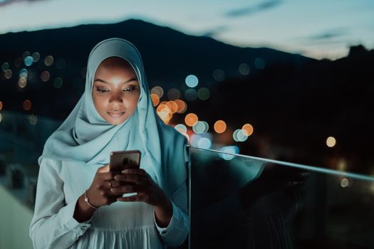 Young Muslim woman wearing scarf veil on urban city street at night texting on a smartphone with bokeh city light in the background. High-quality photo