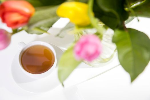 A bouquet of multi-colored tulips in a transparent vase and cup of tea and tea pot on the white table.