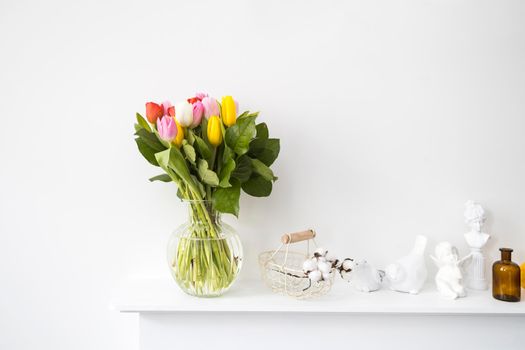 A bouquet of multi-colored tulips, a wicker metal basket with cotton flowers inside. A figurine of a bird, a female bust, two faience birds stand on the panel of a fake hearth. Brown pharmacy bottle.