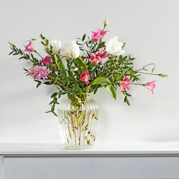 A minimalistic bouquet of white tulips, pink eustoma, hyacinth, eucalyptus in a fluted glass vase on a white panel of an artificial fireplace. Square frame. Empty space.