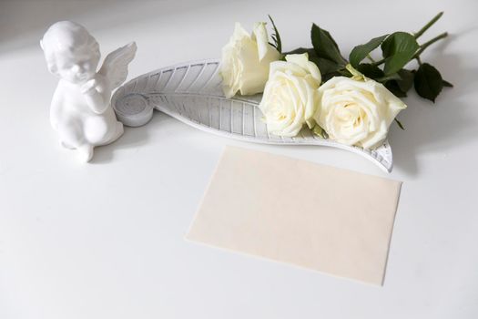 a white faience angel and bunch of roses are on the table. Card