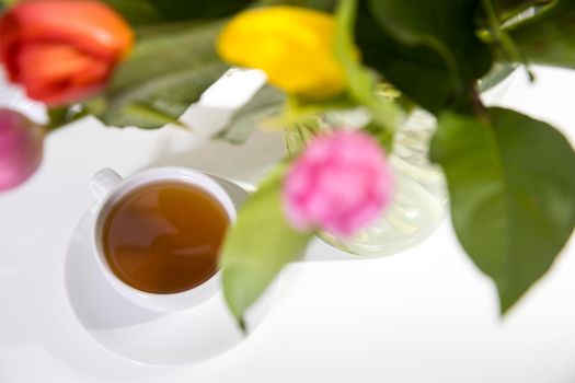 A bouquet of multi-colored tulips in a transparent vase and cup of tea and tea pot on the white table.