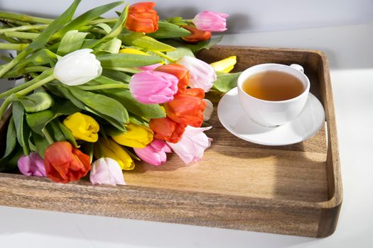 A bouquet of multicolored tulips and a cup of tea are on a wooden tray on the table