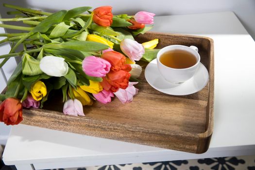 A bouquet of multicolored tulips and a cup of tea are on a wooden tray on the table