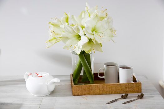 A bouquet of white lily in a glass vase on a table with two tall cups of coffee, a teapot, spoons. Copy space