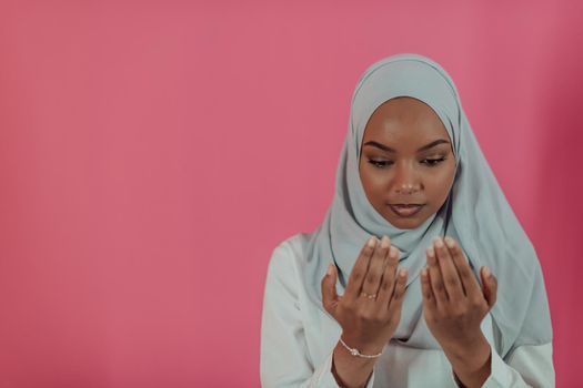 Modern African Muslim woman makes traditional prayer to God, keeps hands in praying gesture, wears traditional white clothes, has serious facial expression, isolated over plastic pink background. High-quality photo