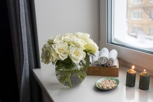 A bouquet of white roses in round glass vase on window with gray curtains. Tray with white small hand towels in spa salon. Green candles with aroma. Plate in the form of green leaf with small stones.