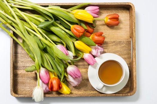 A bouquet of multicolored tulips and a cup of tea are on a wooden tray on the table. Copy space