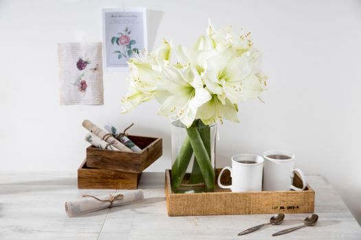 Moscow, Russia - 25 December 2020, A bouquet of white lily in a glass vase on a table with two tall cups of coffee, a teapot, spoons, and a milk jug. Copy space