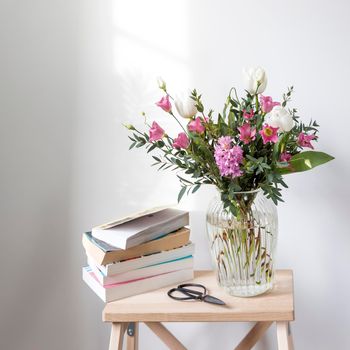A minimalistic bouquet of white tulips, pink eustoma, hyacinth, eucalyptus in a fluted glass vase on a white panel of an artificial fireplace. Empty space. A stack of books and scissors.