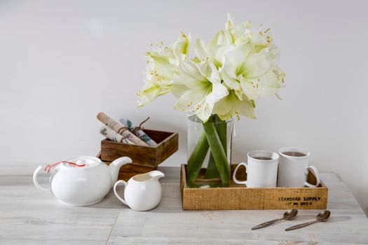 A bouquet of white lily in a glass vase on a table with two tall cups of coffee, a teapot, spoons, and a milk jug. Copy space