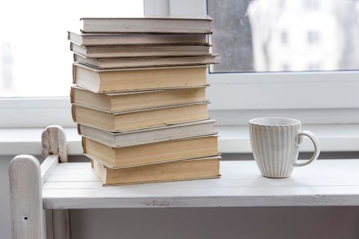 A stack of white-bound books is on the table against the background of the window. The cup of tea is next to books. School education concept.