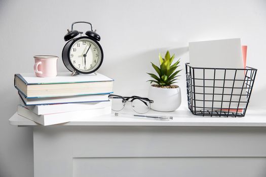 A stack of books, an artificial succulent plant in a ceramic pot, glasses, a metal basket with notebooks, a pen and a cup on a white table surface. Concept - office at home. Online training.