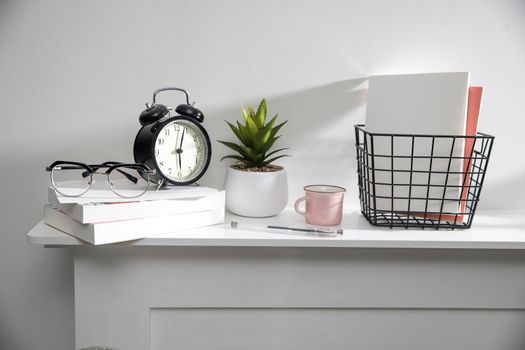 A stack of books, an artificial succulent plant in a ceramic pot, glasses, a metal basket with notebooks, a pen and a cup on a white table surface. Concept - office at home. Online training.