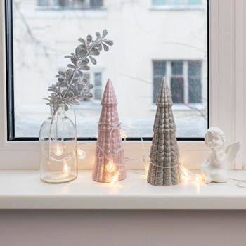 Gray and pink ceramic Christmas trees, a glass bottle with an artificial silver branch and a garland decorate the windowsill for Christmas.