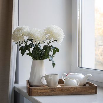 Three white chrysanthemums in a seventies-style fluted vase. Wooden tray with a kettle, milk jug and a cup of tea on the windowsill. Morning breakfast.