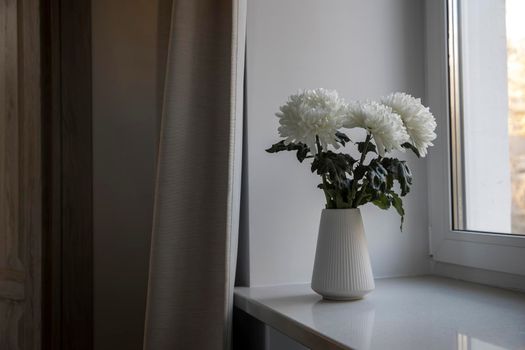 Three white chrysanthemums in a seventies-style fluted vase on the windowsill. The grey curtain