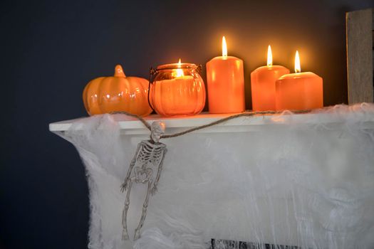 Halloween home decoration. Plastic toy skeletons in a wooden box on a fireplace against a dark blue wall. A garland of skeletons. Cobweb on the dresser. Orange candles and lantern.