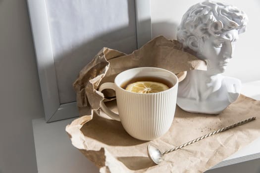 A white corrugated cup with tea and lemon, with a plaster head sculpture of Apollo and a cupronickel teaspoon on a long handle is on brown craft paper on a wooden table.