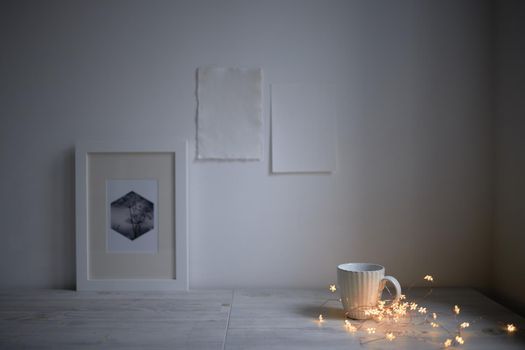 Scandinavian style. Interior Design. A white cup, cozy lights of a luminous garland, a frame for a photo are on the table. Two blank sheets of paper are attached to the wall. Empty space for text