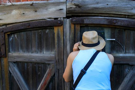 A girl in blue shirt and a straw hat peers through the crack in the door of an old barn