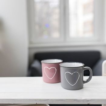 Gray and pink with hearts enamel cups sit on the table in front of the window. Valentine's Day.