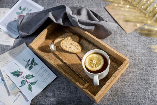 A white cup of tea with lemon, a long cupronickel spoon with a twisted handle and a saucer with three oatmeal cookies for breakfast on a wooden tray, a rag napkin on a gray sofa. Serving