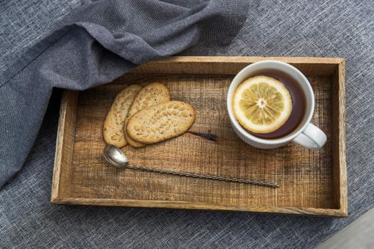 A white cup of tea with lemon, a long cupronickel spoon with a twisted handle and a saucer with three oatmeal cookies for breakfast on a wooden tray, a rag napkin on a gray sofa. Serving