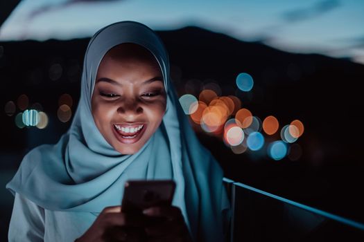 Young Muslim woman wearing scarf veil on urban city street at night texting on a smartphone with bokeh city light in the background. High-quality photo