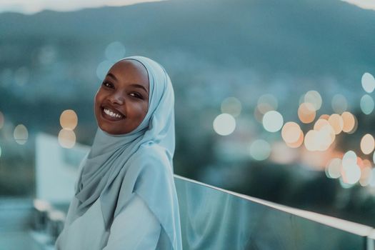 African Muslim woman in the night on a balcony smiling at the camera with city bokeh lights in the background. High-quality photo