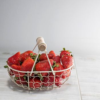 White metal basket is with wooden handle with fresh strawberries on beige table. Square frame