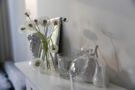 A bunch of fluffy dandelions in a chemical flask, an empty photo frame, a cup of espresso coffee, and a plaster head of David on a white chest of drawers. Copy space