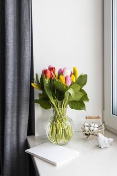 A bouquet of multi-colored tulips in a transparent vase on the windowsill on a holiday. White notebook, wicker metal basket with cotton flowers inside. Bird figurine. Gray curtain.