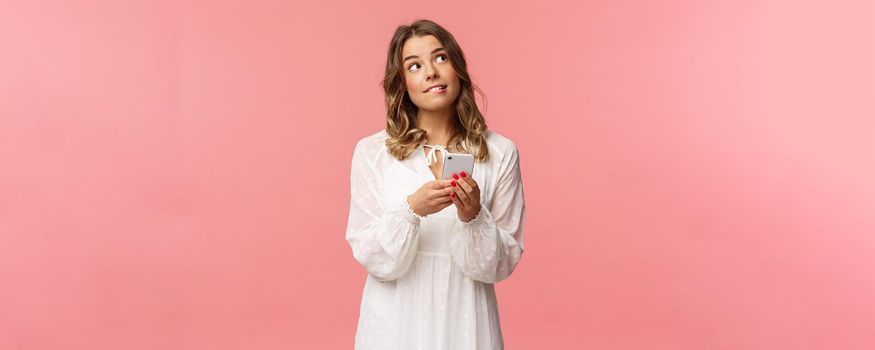 Portrait of dreamy and thoughtful girl thinking what order, using application to make reservation, look up and bite lip pondering, thinking as using mobile phone over pink background.