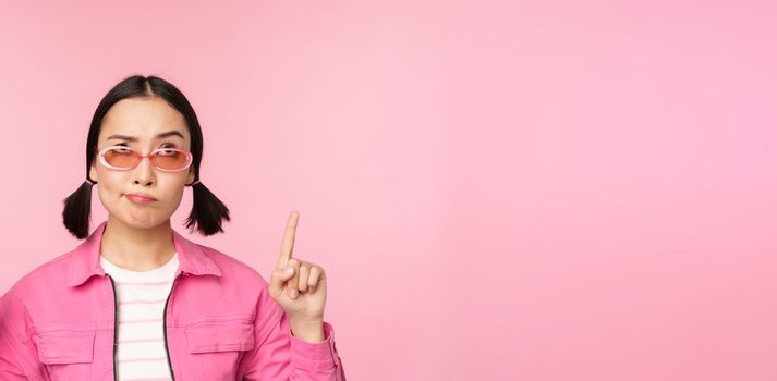Hesitant asian girl, wears sunglasses, frowns and points fingers up, looks with disbelief, skeptical face expression, stands over pink background.