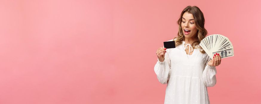 Portrait of rich good-looking blond girl in white dress, holding dollars money and credit card, winning lottery, think how invest, standing happy pink background. Copy space