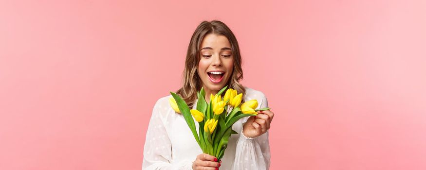 Spring, happiness and celebration concept. Portrait of surprised and pleased happy girl cheerfully looking at bouquet of flowers, smiling amused, receive yellow tulips, stand pink background.