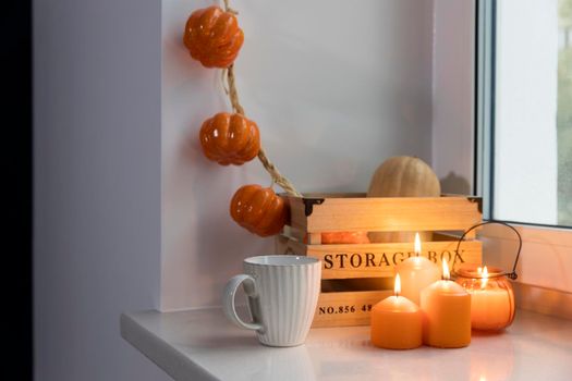 Preparing your home for Halloween. A wooden pumpkin box, a faux pumpkin garland, orange candles, a cup of tea and a candle lantern decorate the window.